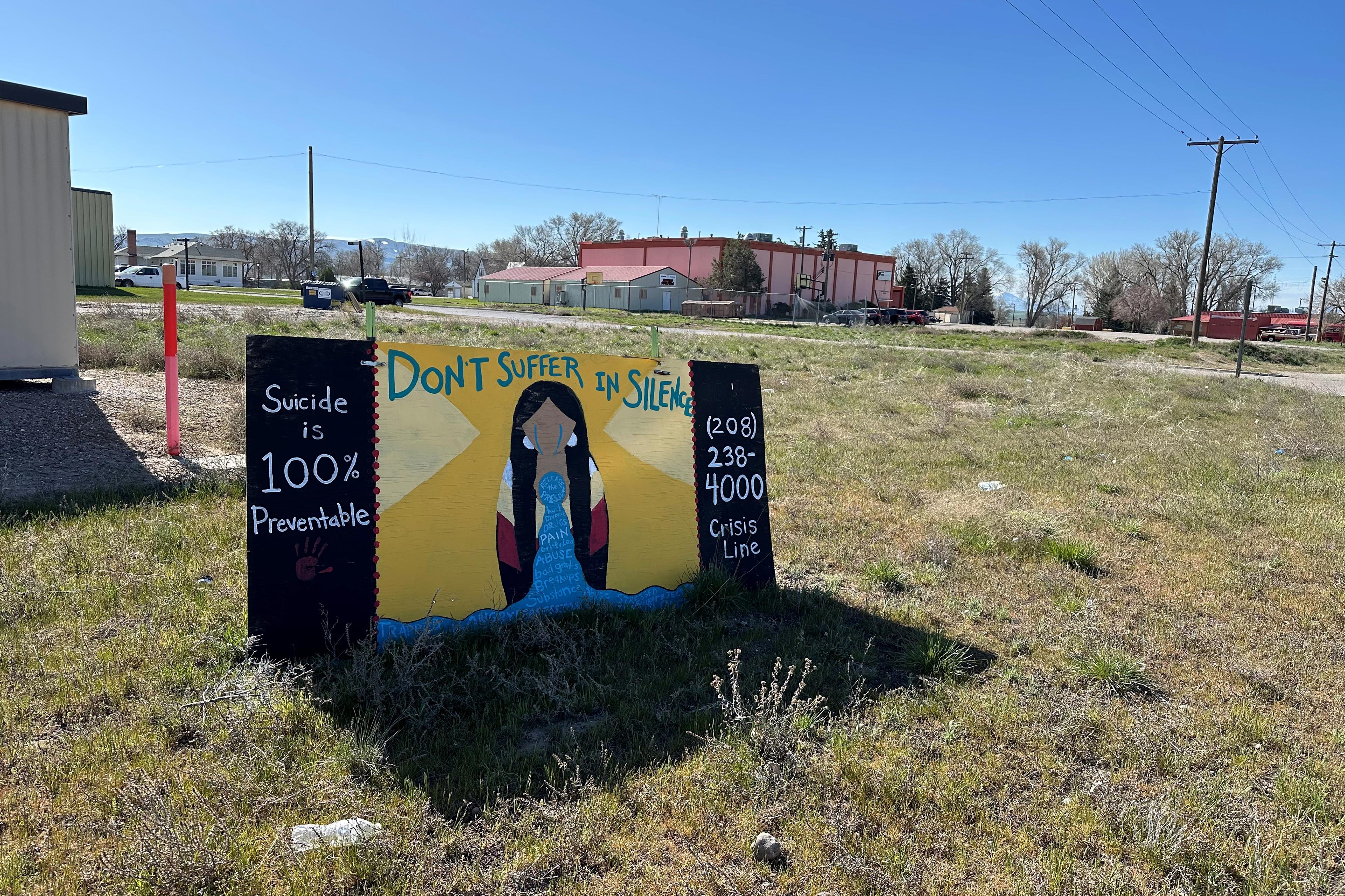 A sign hand-painted with a crying figure in the middle is propped up in the low grass near some buildings reads: "Suicide is 100% Preventable," "Don't suffer in silence," and "208-238-4000 Crisis Line."